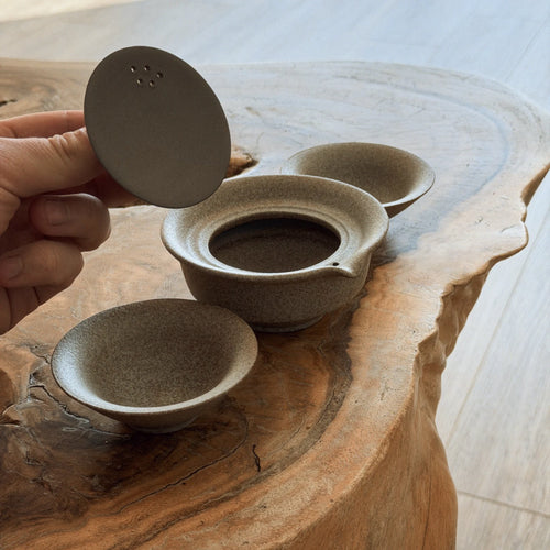 Hand holding open beige ceramic gaiwan lid showing interior, with cup on natural wood slab