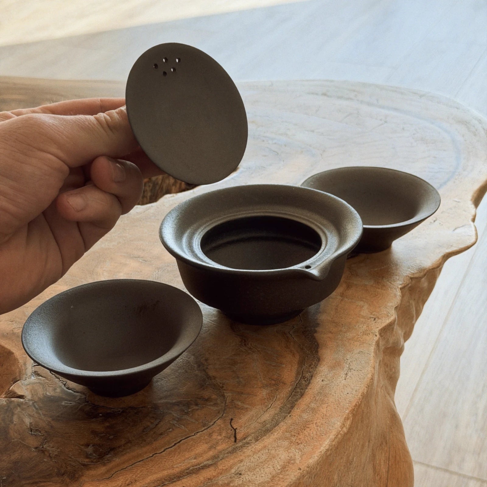 Hand holding open dark ceramic gaiwan lid showing interior, with 2 cups on wooden surface